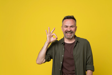 Happy smiling handsome senior bristle man showing sign OK with fingers on one hand isolated over yellow background dressed in green shirt and brown t-shirt