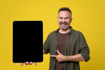 mustached senior man dressed in green shirt pointing with thumb up and index finger to his big tablet pc with black mock-up screen holding in hand isolated on yellow background