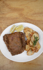 Top view of traditional milanesa, breaded fried steak, with chips and lemon, in a white dish on the wooden table.