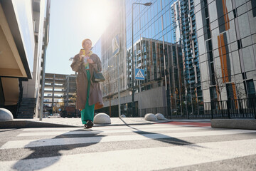 Smiling woman in beige raincoat crosses street at pedestrian crossing