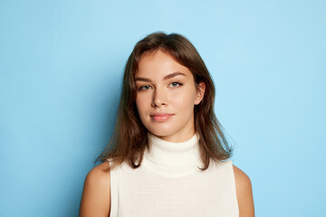 Fototapeta premium Portrait of young beautiful woman posing, looking at camera isolated over blue studio background. Attractive blue eyes