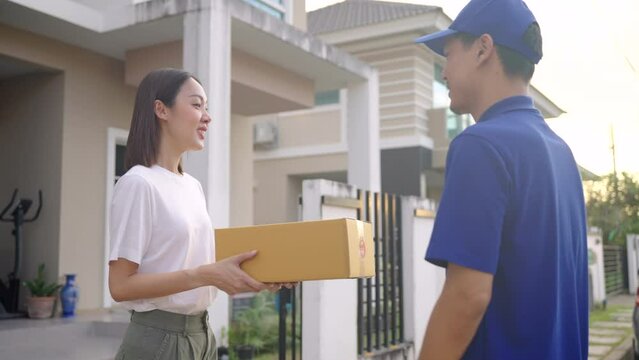 Handsome asian delivery man wearing blue uniform sending a cardboard box to customer in front of the customer's house. 4k resolution.