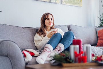 Positive woman in plaid with cup of tea watching movie, TV on sofa at home with christmas decoration atmosphere. Lady wear jumper and warm socks. Cozy and comfortable winter concept. Selective focus.