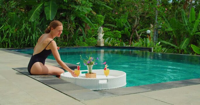 Multiracial woman in black swimsuit drinking non-alcoholic orange cocktail from straw basket. Relaxation girl sitting by the pool water, smiling. Tourist enjoying vacation. Rainforest.