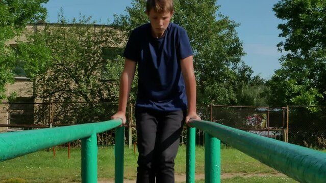  A boy in a blue T-shirt is swinging on the gymnastic bars in the street. Handsome boy 14 years old doing exercises on the gymnastic uneven bars.High quality 4k footage