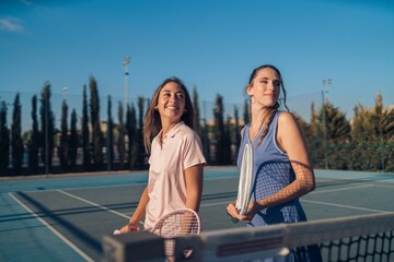 Pair of young female friends with blue and pink athleisure and matching tennis rackets on a court