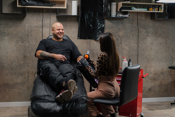 Naklejka na ściany i meble Woman tattoo artist makes a tattoo to a man on his arm, tattoo studio