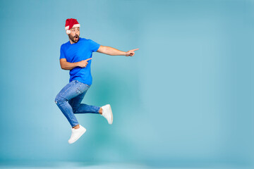 Fototapeta na wymiar Full length portrait of a cheerful young man jumping in a blue t-shirt and a New Year's hat, pointing his finger to the side on a blue background