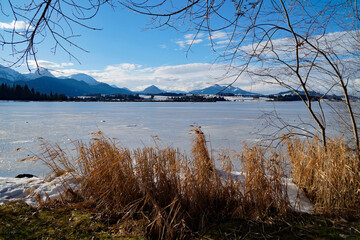 wintery frozen over alpine lake Hopfensee covered with ice with the snowy Bavarian Alps in the background on a sunny but very windy and cold December day, Germany