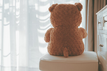 Brown teddy bear sitting alone on white cotton chair with sunlight from the door or window in front of house, Cinematic tone style vintage, Lonely and waiting for lover or family concept.