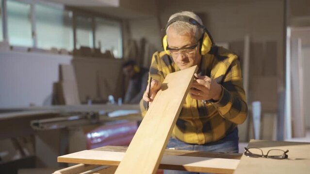 Adult carpenter man working in carpentry workshop measures wood with meter.
Male carpenter measuring dusty wood with meter and marking with pencil.
