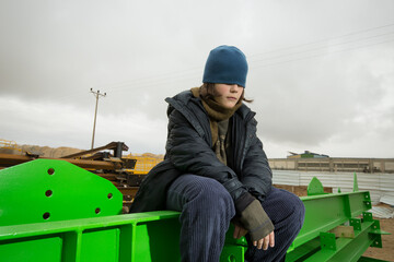 cool stylish caucasian boy with the blue hat on his face covering his eyes in a coat and hat with a scarf posingon green metal structures in an industrial area against a cloudy greysky on a rainy day