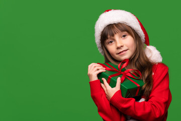 A little girl holds a gift on a green isolated background. A child in a Santa Claus hat and a warm sweater holds a gift box.