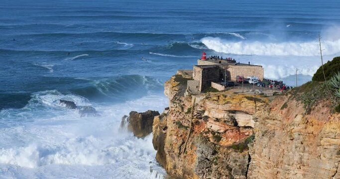 People watching the big giant waves crashing near the Fort of Nazare Lighthouse in Nazare, Portugal. Biggest waves in the world. Touristic destination for surfing.