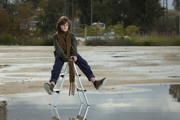 cool stylish brown-haired caucasian boy with a long scarf and a sweater posing on a ladder in a puddle against the background of trees on a rainy day. fashion magazine concept