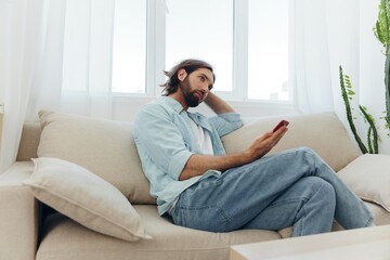 A man with a beard sits on the couch during the day and smiles at home and looks at his phone relaxing on his day off, life online on his phone texting with friends