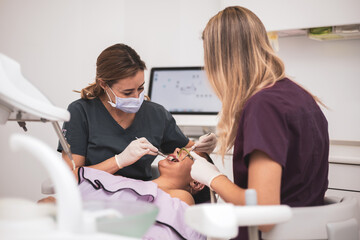 A female dentist and her assistant examine a patient's teeth in the dentist's chair.The hygienist uses an electrical device or an air turbine.