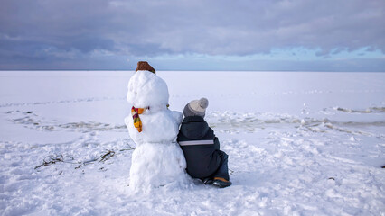 Seven years boy in knitted winter hat sits alone with snowman and looks forward. Loneliness and a friend made of snow. Winter mood on a cloudy day. Banner format