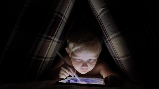 A little boy draws with a stylus on an electronic tablet in a hut made of pillows in the dark. The concept of early development of children. The concept of the presence of gadgets in children's lives