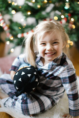 toddler girl wearing plaid pajamas cuddling with her stuffed animal in front of the Christmas tree
