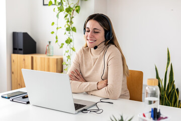 Young adult business woman with headset working on laptop having a video call sitting at home office