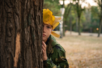 Beautiful teenage boy with crown of yellow leaves playing in the park and hiding behind the tree