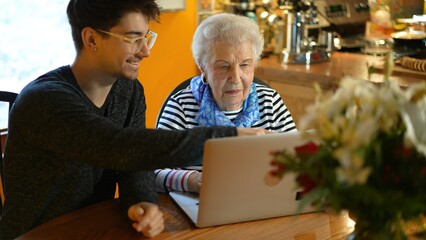 Senior elderly woman and teen grandson happy and smiling learning on laptop computer in dining room.