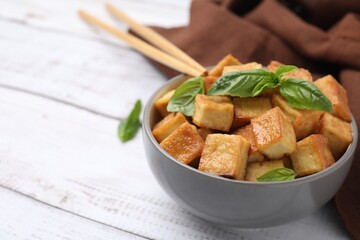 Bowl with delicious fried tofu and basil on white wooden table, closeup. Space for text