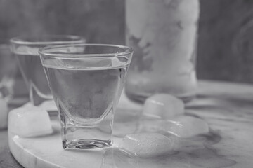 Shot glasses and bottle of vodka with ice cubes on marble board, closeup
