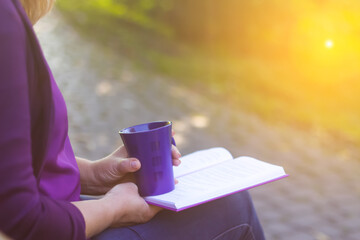 Defocus of woman hand holding and reading a book with holding purple mug cup of hot coffee. A woman sits near a tree in an autumn forest and holds a book and a cup in her hands. Out of focus