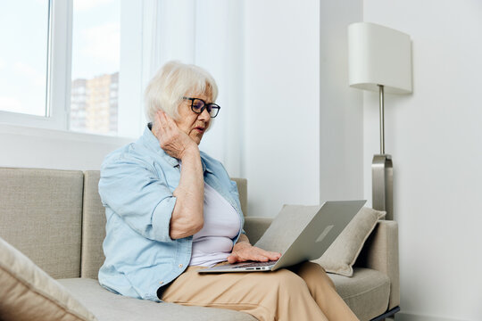 a sad elderly woman is sitting on a sofa in a bright apartment and using a laptop is looking for information on the Internet, holding her hand near her head in frustration, looking with regret - Powered by Adobe