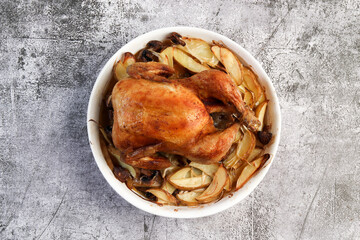Whole roast chicken with potatoes and onions in a white baking dish on a dark grey background. Top view, flat lay