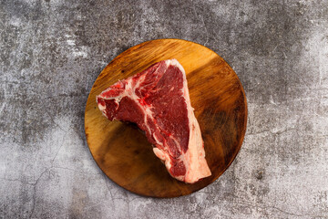 Raw t-bone beef steak on a round wooden cutting board on a dark grey background. Top view, flat lay