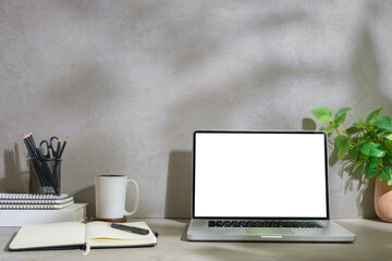 Laptop computer with empty display, notepad, coffee cup, stationery and houseplant on grey table.