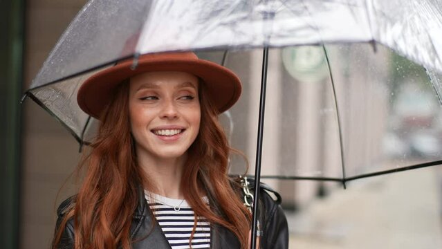 Portrait of cheerful redhead young woman wearing stylish hat holding transparent umbrella standing on gray city street in rainy weather rain, smiling looking at camera. Shooting in slow motion.
