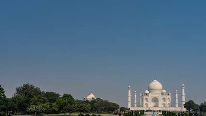 Beautiful ancient mausoleum of the Taj Mahal against the blue sky. Minarets, towers, domes, arches made of white marble. Green vegetation in the park. A lot of people are sightseeing. Copy space.