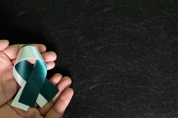 Top view of human hand palm holding teal and white ribbon in dark black background. Cervical cancer...