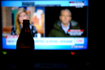 Drinking beer while watching the news. A bottle of beer is standing in the dark, in the background is a bright screen with the evening news