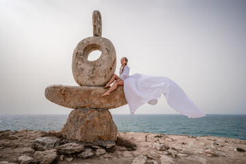 Fototapeta na wymiar A woman sits on a stone sculpture made of large stones. She is dressed in a white long dress, against the backdrop of the sea and sky. The dress develops in the wind.