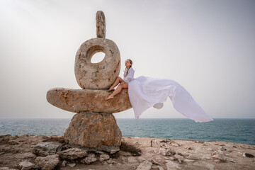 A woman sits on a stone sculpture made of large stones. She is dressed in a white long dress,...