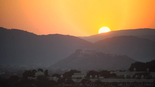 Evening dusk sunset shot showing the orange sun setting behind the layers of aravalli range with trees and old heritage buildings in the foreground in udaipur rajasthan India