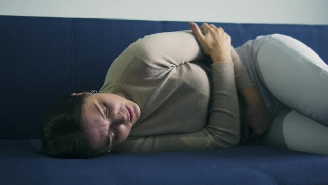 Deeply upset brunette woman lying down on sofa and touching her belly, feeling unbearable stomach ache, suffering dysmenorrhea, painful menstruation and abdominal cramps, miscarriage, abortion