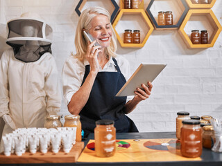 Small business woman, tablet and phone call in honey store for consulting, marketing or advertising in retail. Happy elderly female business owner working in a organic shop on phone with touchscreen