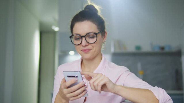 Smiling woman in eyeglasses sitting on sofa and scrolling web-pages on smartphone, doing shopping online, checking on-line stores with discounts and sales, black Friday sale