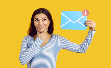 Hmm, interesting, what's new in mail. Studio shot of happy beautiful young woman standing on yellow color background, looking at blue paper envelope with red message notification, thinking and smiling - Powered by Adobe