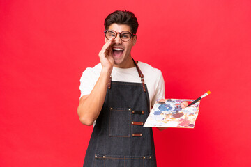 Young artist caucasian man holding a palette isolated on red background shouting with mouth wide...