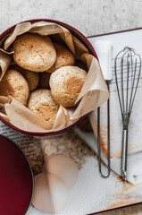 Homemade cookies on baking paper in a round metal container with a burgundy lid standing on an open cookbook, with a whisk and a silicone scapula on the background of the ceramic tile, top view.