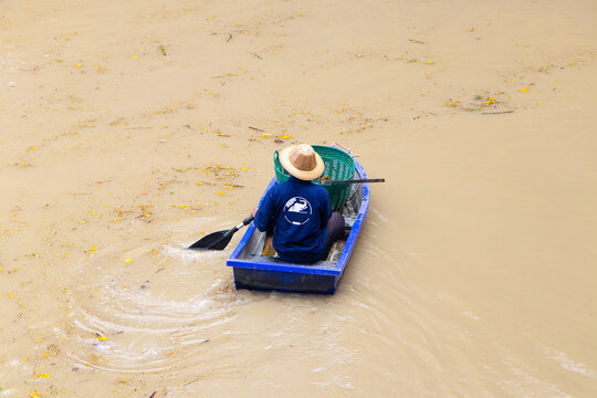 Male old Boatman wearing blue local dress rowing a boat with a bamboo in a dirty freshwater canal, orange-red at Thailand. during the day under the bright sunlight. to find fish for dinner for family.