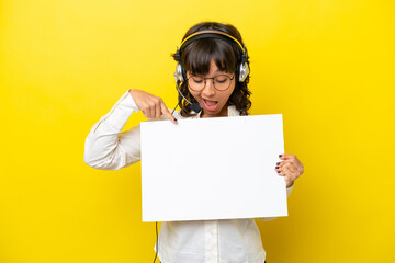 Telemarketer latin woman working with a headset isolated on yellow background holding an empty placard with happy expression and pointing it