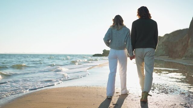 Back view of lovely couple holding hands and walking along the beach by the sea